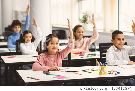 Return Back To School. Multiciltural diverse group of happy small elementary schoolchildren sitting at desks in classroom, raising hands up for an answer, learning with pleasure. Social distance 127412874