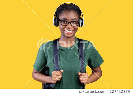 Happy African Female Student Posing Wearing Wireless Headphones Listening To Educational Podcast Online Standing On Yellow Studio Background, Smiling To Camera. University Student's Portrait Concept Happy African Female Student Posing Wearing Wireless Headphones Listening To Educational Podcast Online Standing On Yellow Studio Background, Smiling To Camera. University Student's Portrait Concept 127412879