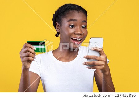Mobile Shopping. Excited Black Woman Holds Credit Card And Smartphone, Shops Online Via Application Standing Over Yellow Studio Background. Excited Customer, Shopping App And Cashback 127412880