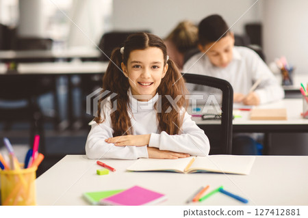 First Day At School. Portrait of smiling diligent small girl with ponytails sitting at table in classroom at elementary school with group of classmates in background and posing, looking at camera 127412881