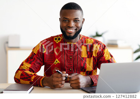 Handsome African Freelancer Guy In Traditional Costume Sitting At Desk At Home Office, Working On Laptop Computer And Smiling At Camera, Black Man Enjoying Remote Work And Freelance Career, Free Space Handsome African Freelancer Guy In Traditional Costume Sitting At Desk At Home Office, Working On Laptop Computer And Smiling At Camera, Black Man Enjoying Remote Work And Freelance Career, Free Space 127412882