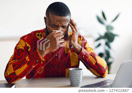 Sick African Man In Traditional Shirt Feeling Unwell While Working In Office, Sitting Desk With Laptop And Blowing Nose In Paper Napkin. Ill Black Guy Suffering Coronavirus Symptoms, Free Space 127412887