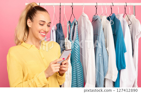 Portrait of smiling fashion blogger influencer holding and using her smartphone, texting sms, checking social media, writing post or making purchases in online store standing near clothing rail 127412890