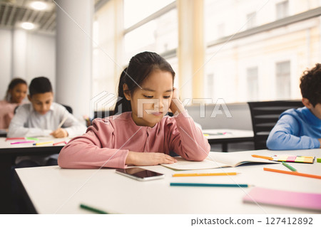 Portrait of focused small asian girl sitting at table in classroom at primary school or kindergarten, reading task in notebook. Reopening and return back to school after coronavirus quarantine Portrait of focused small asian girl sitting at table in classroom at primary school or kindergarten, reading task in notebook. Reopening and return back to school after coronavirus quarantine 127412892