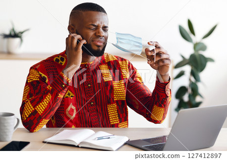 Displeased African Man Taking Off Mask At Work To Scratch Beard, Suffering From Itchy Face Skin, Stressed Black Male In Traditional Clothes Having Allergy, Sitting At Workplace In Office, Free Space 127412897