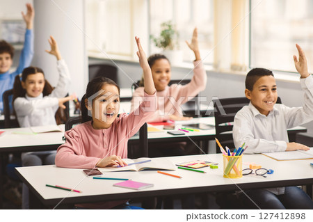 Academic Concept. Diverse group of multiethnic smart junior schoolchildren sitting at desks in classroom and raising hands for an answer, studying with pleasure. Positive young elementary students 127412898
