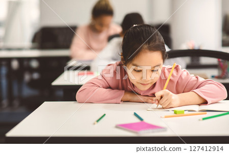Tuition Concept. Portrait of concentrated small asian girl sitting at table in classroom, writing or drawing in notebook, doing homework or taking notes during lesson, selective focus, free copy space 127412914