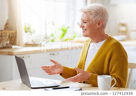 Cheerful senior woman having online class from home, talking to teacher or lecturer, sitting on kitchen in front of modern laptop. Positive elderly lady having video chat, using notebook, copy space 127412938