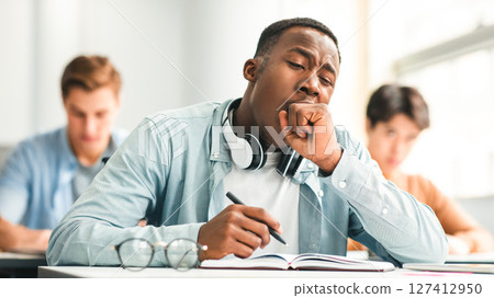 Tired black student yawning sitting at desk in classroom. African American youth exhausted from getting ready for test or writing coursework, feeling sleepy during lecture in highschool. Lack of sleep 127412950
