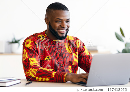 Successful Black Businessman In African Traditional Clothes Sitting At Workplace In Office, Happy Male Entrepreneur In Ethnic Shirt Working With Laptop Computer At Desk, Typing On Keyboard And Smiling 127412953