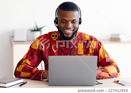 Distance Education. Smiling Black Man In African Shirt And Headset Watching Webinar On Laptop At Home, Having Online Lesson, Positive Millennial Guy In Ethnic Costume Sitting At Desk With Computer Distance Education. Smiling Black Man In African Shirt And Headset Watching Webinar On Laptop At Home, Having Online Lesson, Positive Millennial Guy In Ethnic Costume Sitting At Desk With Computer 127412958