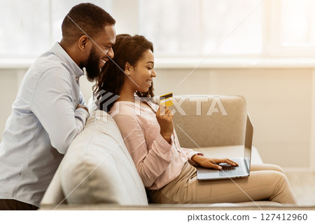 Online Shopping From Home, E-commerce Concept. Happy young african american couple sitting on sofa at home and buying clothes or products online, paying with credit card using laptop Online Shopping From Home, E-commerce Concept. Happy young african american couple sitting on sofa at home and buying clothes or products online, paying with credit card using laptop 127412960
