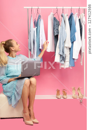 Fashion Designer. Vertical portrait of happy stylish lady sitting in closet on pouffe with computer on lap near rack with clothes and shoes, choosing outfit, isolated on pastel pink studio background 127412986