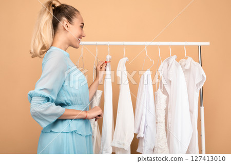 Choosing New Outfit. Cheerful young woman shopping in store, searching for clothes, consumer looking through hangers with stylish white shirts and dresses on clothing rail, standing at wardrobe 127413010