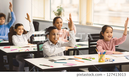 Ready For An Answer. Diverse group of multicultural cheerful primary schoolkids sitting at table in classroom and raising hands, studying and learning. Portrait of positive little elementary students 127413013