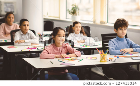 Back To School. Portrait of focused multicultiral pupils sitting with folded arms at desks with social distance, listening to teacher. Diverse junior students working diligently and learning new stuff 127413014
