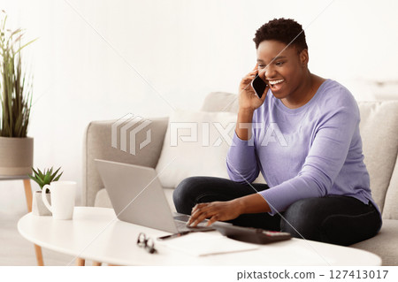 Informal Call. Emotional young curvy african american woman talking on her smartphone, sitting on the couch, using laptop, typing on computer keyboard. Copy space. Communication Concept. Informal Call. Emotional young curvy african american woman talking on her smartphone, sitting on the couch, using laptop, typing on computer keyboard. Copy space. Communication Concept. 127413017