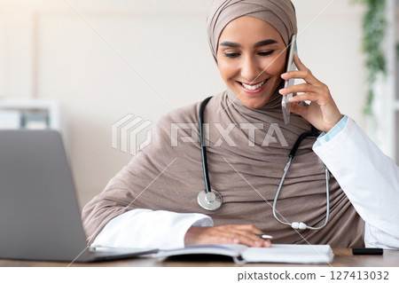 Smiling young lady in hijab doctor talking on mobile phone and checking her notebook, making appointment, sitting at workdesk in front of laptop at clinic, copy space. Remote healthcare, telehealth 127413032