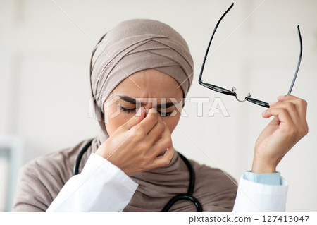 Closeup of female doctor woman in hijab removing glasses and rubbing nose bridge, suffering from tired eyes syndrome, clinic interior. Eye fatigue, symptoms and treatments concept Closeup of female doctor woman in hijab removing glasses and rubbing nose bridge, suffering from tired eyes syndrome, clinic interior. Eye fatigue, symptoms and treatments concept 127413047