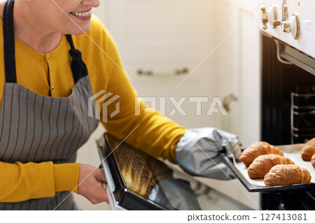 Closeup of smiling elderly woman baking pastry at home, taking delicious croissants out of oven, copy space. Joyful senior lady in apron opening oven, checking her pastry, cozy kitchen interior 127413081