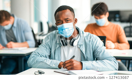 New Normal Education. Portrait of black male student sitting at desk, writing examination or taking notes in textbook during lecture, afro guy in headset and surgical mask posing and looking at camera New Normal Education. Portrait of black male student sitting at desk, writing examination or taking notes in textbook during lecture, afro guy in headset and surgical mask posing and looking at camera 127413087