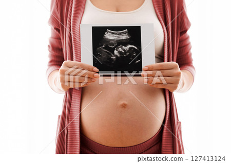 Unrecognizable pregnant woman holding her baby sonography shot near naked belly while standing against window at home, expectant mother enjoying first photo of her unborn child, closeup image, cropped Unrecognizable pregnant woman holding her baby sonography shot near naked belly while standing against window at home, expectant mother enjoying first photo of her unborn child, closeup image, cropped 127413124