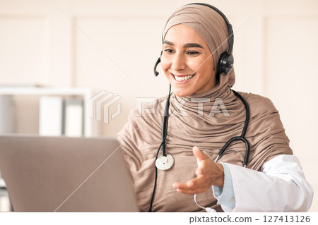 Cheerful arab female doctor working online in the clinic, using headset and laptop. Muslim woman therapist having video conference with her patient during isolation, hospital interior, copy space 127413126