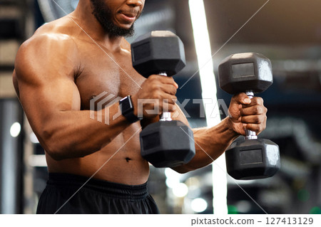 Hot black guy exercising with two dumbbells, working on his muscular biceps, looking at copy space. Concentrated handsome african american shirtless bodybuilder making work out with barbells at gym 127413129