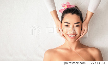 Facial Massage. Attractive young asian lady getting face treatment while relaxing in spa, enjoying beauty procedure, professional beautician touching smiling korean woman's face, top view, copy space 127413131