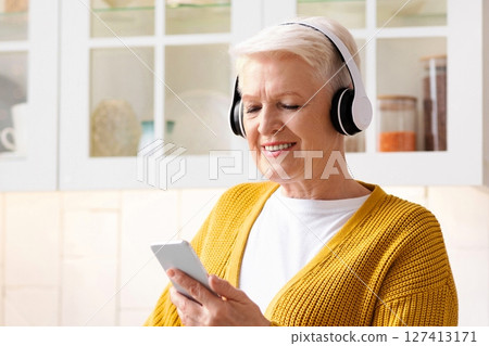 Happy senior grey-haired woman listening to music on smartphone at home, using wireless headphones. Attractive grandmother with mobile phone and headset, kitchen interior, copy space 127413171