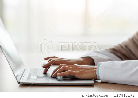Cropped of woman doctor having online training, sitting in her cabinet, using laptop, typing on keyboard, side view, copy space. Unrecognizable lady physician working with computer, clinic interior 127413172