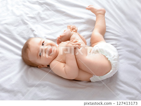 Beautiful caucasian baby laying on bed, playing with feet and smiling at camera, top view, copy space. Adorable toddler in diaper having fun alone, lying on white, laughing. Toddler, baby, infant 127413173