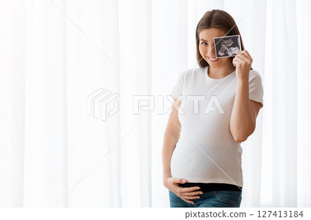 Happy Pregnant Woman With Ultrasound Scan Image Posing Near Window At Home, Cheerful Expecting Lady Embracing Her Belly And Smiling At Camera, Enjoying Healthy Pregnancy And Prenatal Time, Copy Space 127413184