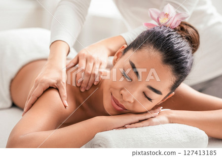 Relaxed asian woman enjoying back and shouders massage in spa. Professional female masseur therapist doing wellness beauty treatment to attractive korean lady with flower in hair, closeup shot 127413185