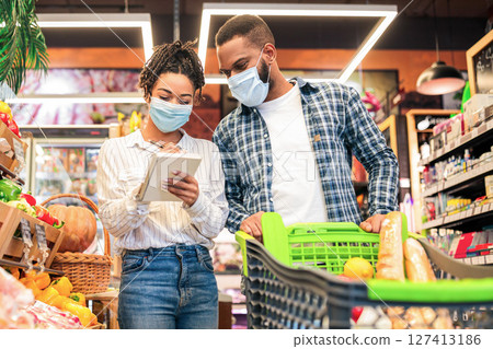 Family In Supermarket. Black Couple Buying Food With Shopping Cart, Wearing Face Masks In Grocery Store Indoors. Shop Customers With Trolley Full Of Goods. Sales And Discounts, Consumerism Concept Family In Supermarket. Black Couple Buying Food With Shopping Cart, Wearing Face Masks In Grocery Store Indoors. Shop Customers With Trolley Full Of Goods. Sales And Discounts, Consumerism Concept 127413186