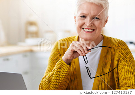 Online education, courses, webinar concept. Cheerful senior woman sitting in front of laptop in kitchen, holding glasses and smiling, drinking tea, enjoying studying online from home, copy space 127413187
