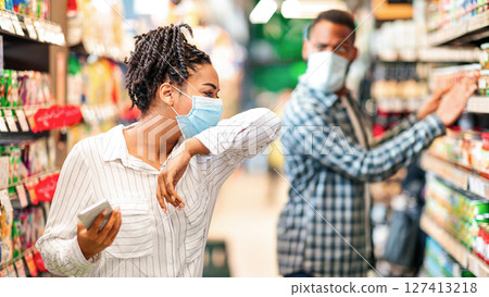 Sick African Woman In Supermarket Coughing In Elbow Wearing Mask Doing Grocery Shopping Buying Food Indoor. Shop Safe, Coronavirus Covid-19 Pandemic Outbreak, Social Distance In Public Places. Sick African Woman In Supermarket Coughing In Elbow Wearing Mask Doing Grocery Shopping Buying Food Indoor. Shop Safe, Coronavirus Covid-19 Pandemic Outbreak, Social Distance In Public Places. 127413218