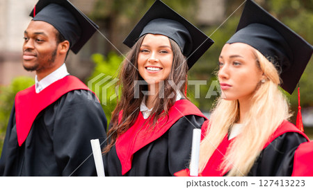Attractive young brunette lady standing among international group of students and smiling, wearing graduation costumes and holding diplomas, multiracial graduates having graduation ceremony, closeup 127413223
