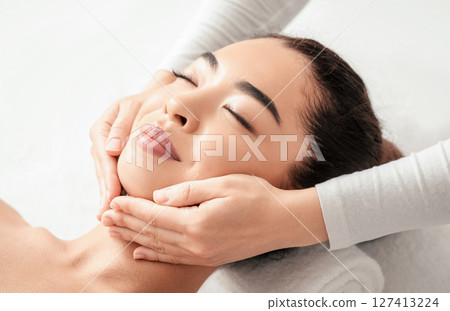 Face Massage. Closeup of attractive asian woman getting beauty treatment at spa salon, relaxed young korean lady enjoying skin and body care, lying on massage table with closed eyes, free space 127413224