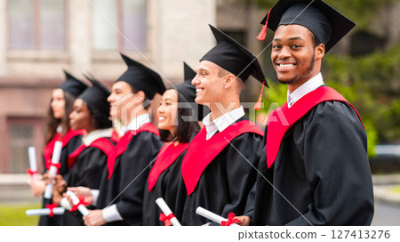 Diverse international students with diplomas attending graduation ceremony, happy multiracial group of students in graduation caps and robes cheerfully standing in a row at university campus 127413276