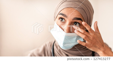 Avoid face touching, Coronavirus prevention, Doctors protection during COVID-19 pandemic. Overworked female doctor in protective face mask and hijab touching her eyes, closeup portrait Avoid face touching, Coronavirus prevention, Doctors protection during COVID-19 pandemic. Overworked female doctor in protective face mask and hijab touching her eyes, closeup portrait 127413277