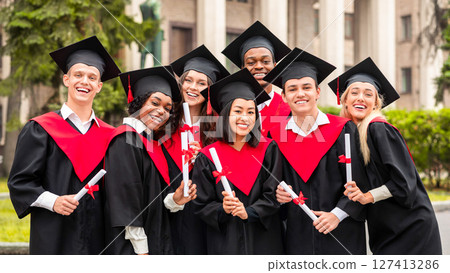 Cheerful multiracial students in graduation robes and hats celebrating graduation at university campus, posing together outdoors, smiling at camera, closeup group portrait 127413286