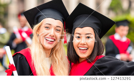 Two cheerful multiracial young ladies girlfriends in graduation clothes taking selfie, happy female students showing diplomas and smiling at camera, closeup portrait over university campus Two cheerful multiracial young ladies girlfriends in graduation clothes taking selfie, happy female students showing diplomas and smiling at camera, closeup portrait over university campus 127413291