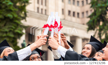 Happy multiracial group of students in graduation caps and robes cheerfully posing at university campus, joining together their diplomas, celebrating graduation, closeup, copy space Happy multiracial group of students in graduation caps and robes cheerfully posing at university campus, joining together their diplomas, celebrating graduation, closeup, copy space 127413296