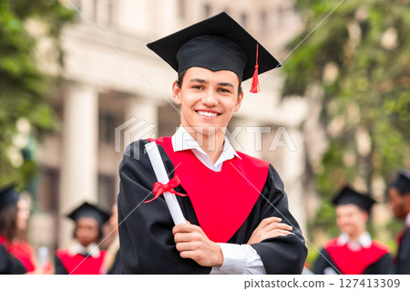 Cheerful guy in graduation costume showing his diploma and smiling at camera, happy male student posing over international group of students at university campus, copy space 127413309