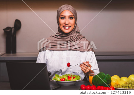 Arab Lady Eating Vegetable Salad For Dinner Sitting At Laptop Computer In Modern Kitchen At Home, Smiling To Camera. Dieting Blog, Healthy Food Online Recipes And Weight Loss Nutrition Concept 127413319