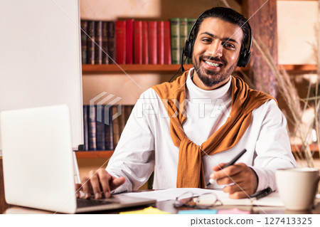 E-Teaching. Smiling Arab Teacher Guy In Headphones Sitting At Laptop Posing Looking At Camera In Classroom. Online Teaching, Modern Distance Education And Remote E-Learning Concept 127413325