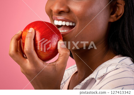Healthy teeth concept. Pretty black woman with beautiful smile biting fresh red apple on pink studio background. Millennial African American lady with healthy dentals eating ripe fruit 127413342