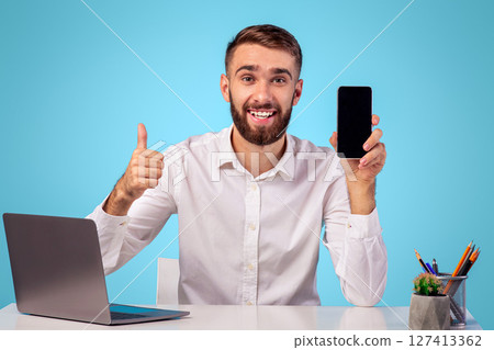 Positive young office employer showing smartphone with empty screen and showing thumb up gesture at workplace, mockup for mobile app or website design. CEO recommending online business service 127413362