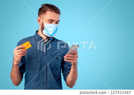 Contactless shopping during covid isolation. Portrait of cool millennial guy in face mask holding smartphone and credit card over blue studio background. Young man buying goods in online store Contactless shopping during covid isolation. Portrait of cool millennial guy in face mask holding smartphone and credit card over blue studio background. Young man buying goods in online store 127413367